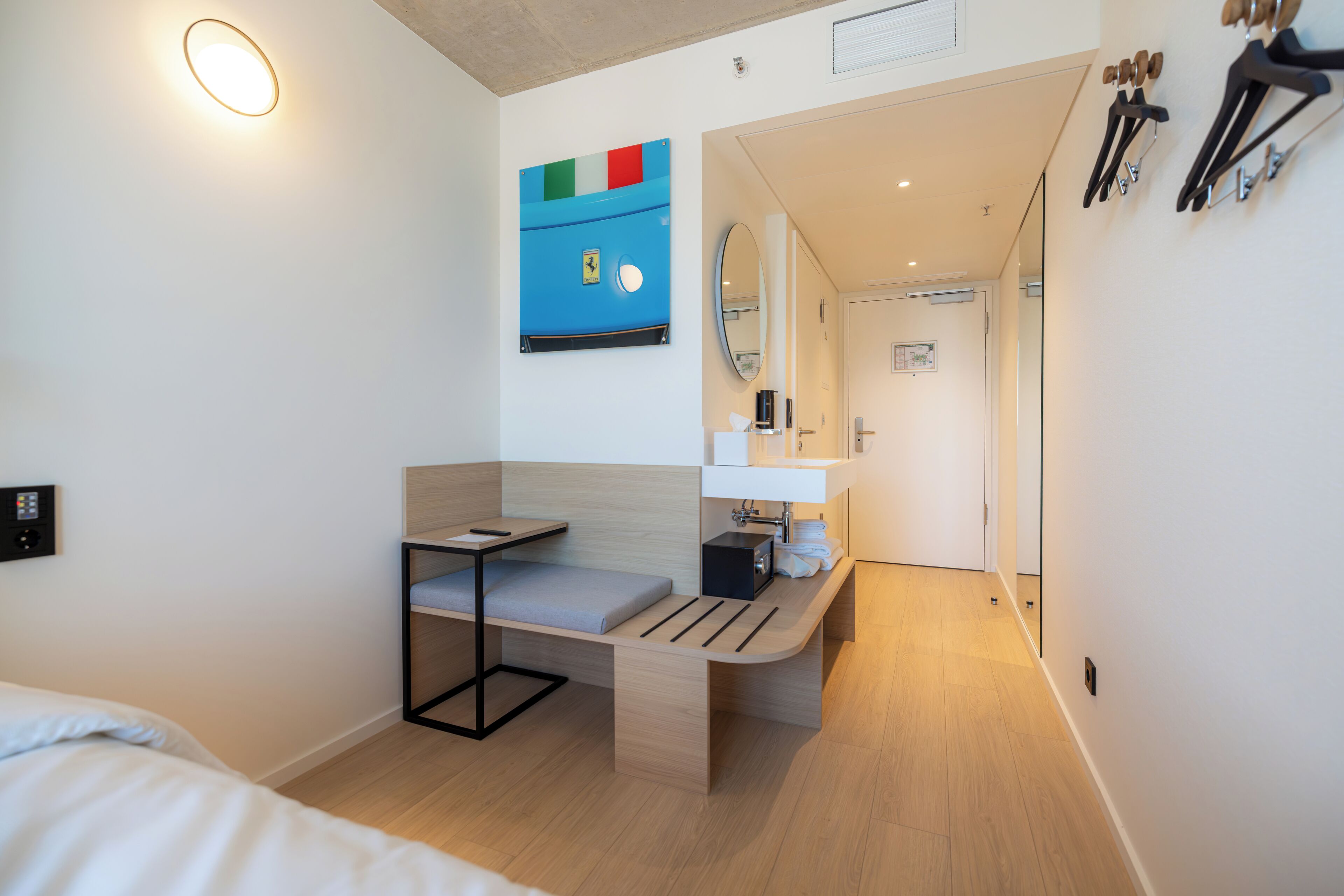 A modern hotel room with a bed, a desk, a bench, a mirror, and a bathroom visible through an open door.