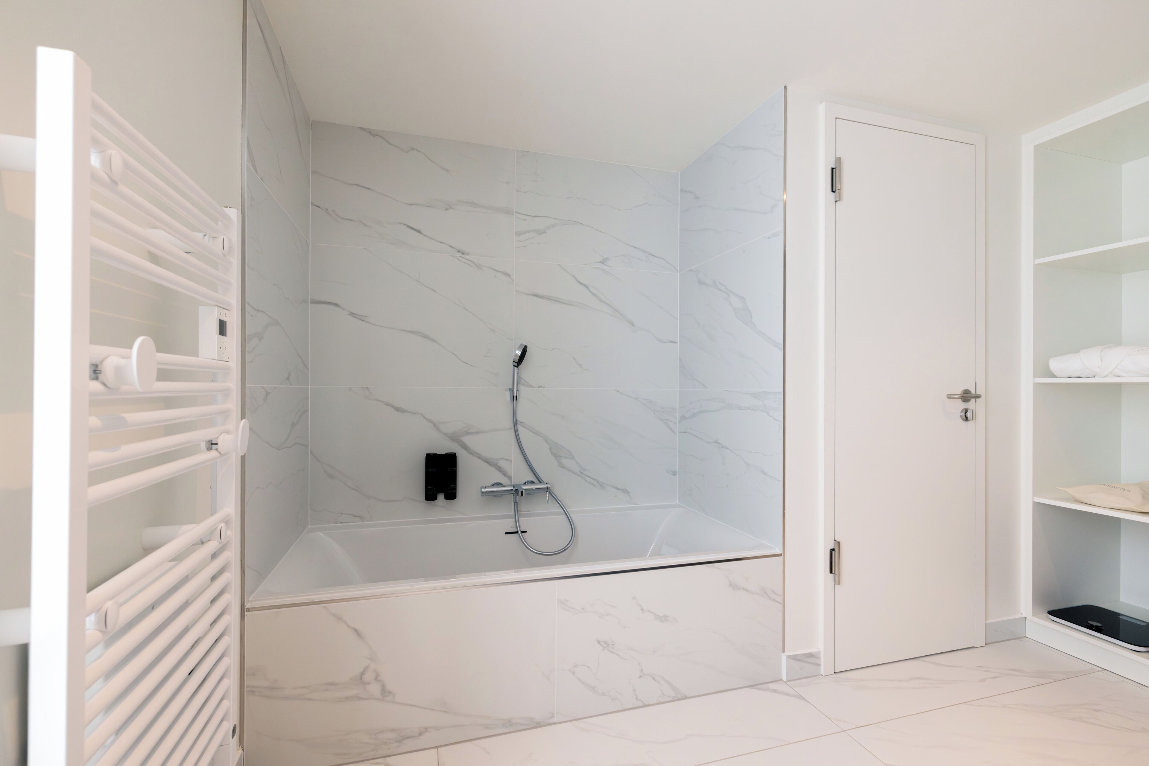Un baño moderno con ducha a ras de suelo, baldosas de mármol, una puerta blanca y estanterías empotradas.