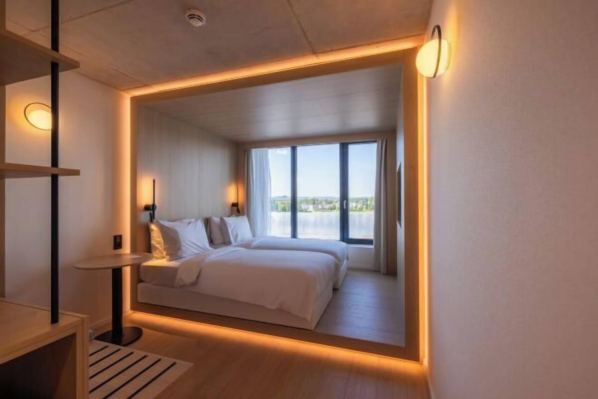 Un dormitorio moderno con una cama grande, una mesita pequeña y vistas al exterior a través de un ventanal amplio.