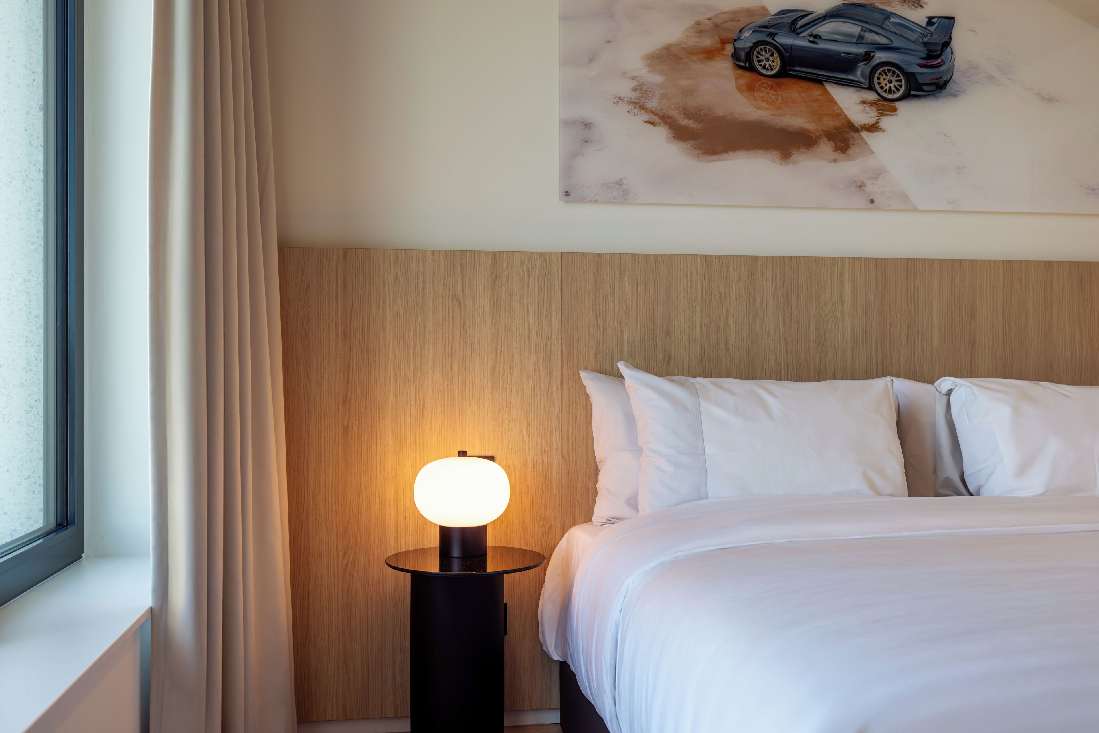 A hotel room with a bed, white bedding, a nightstand with a lamp, a painting of a car on the wall, and a wooden headboard.