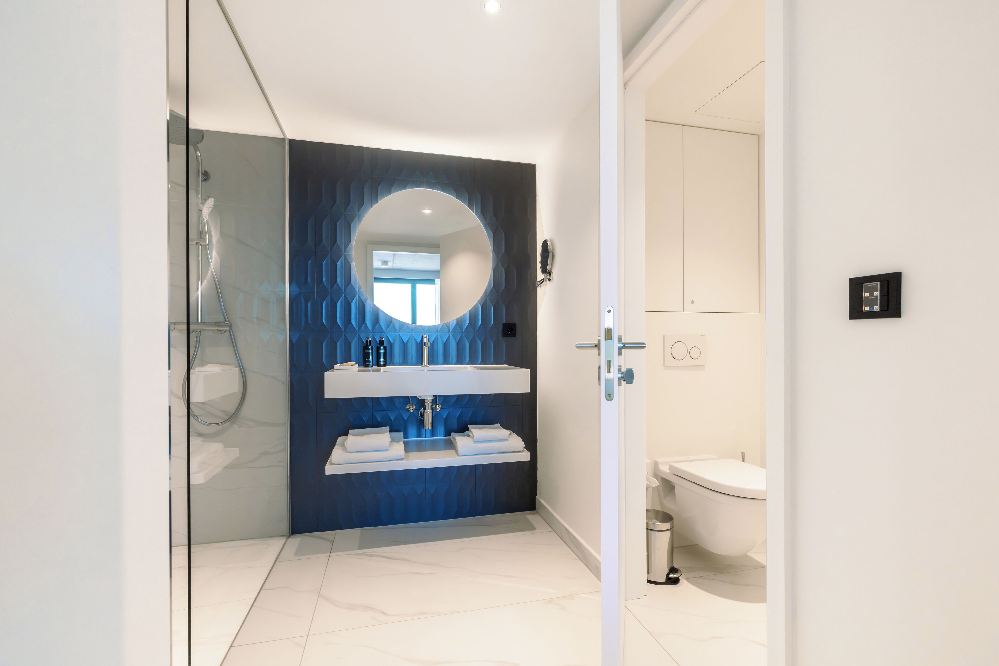 Un baño moderno con cabina de ducha de vidrio, un espejo redondo y un lavabo blanco con una pared de acento azul.