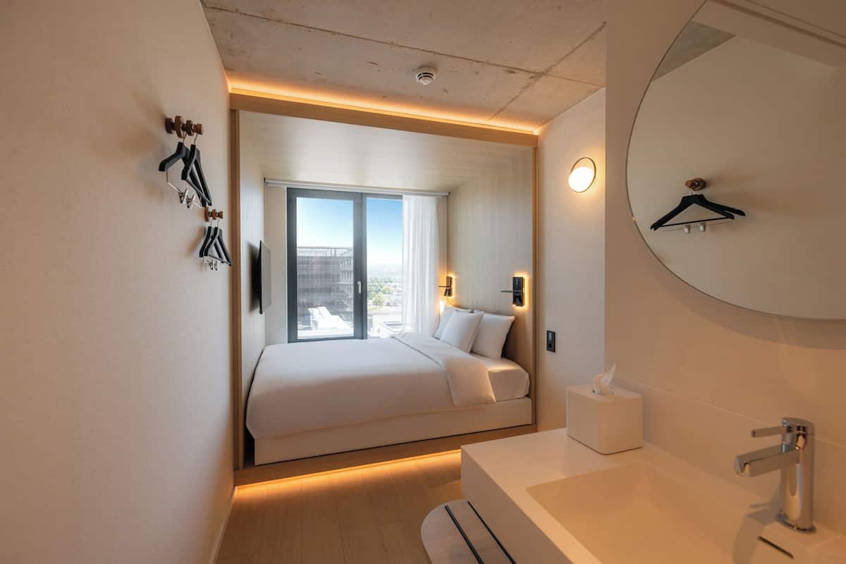 Habitación de hotel moderna con cama, mesita de noche, espejo y vistas a la ciudad.