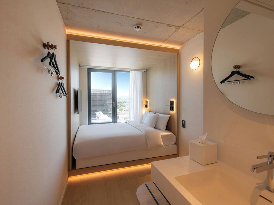 Habitación de hotel moderna con una cama grande, una mesita de noche, un espejo y vistas a la ciudad a través del ventanal.