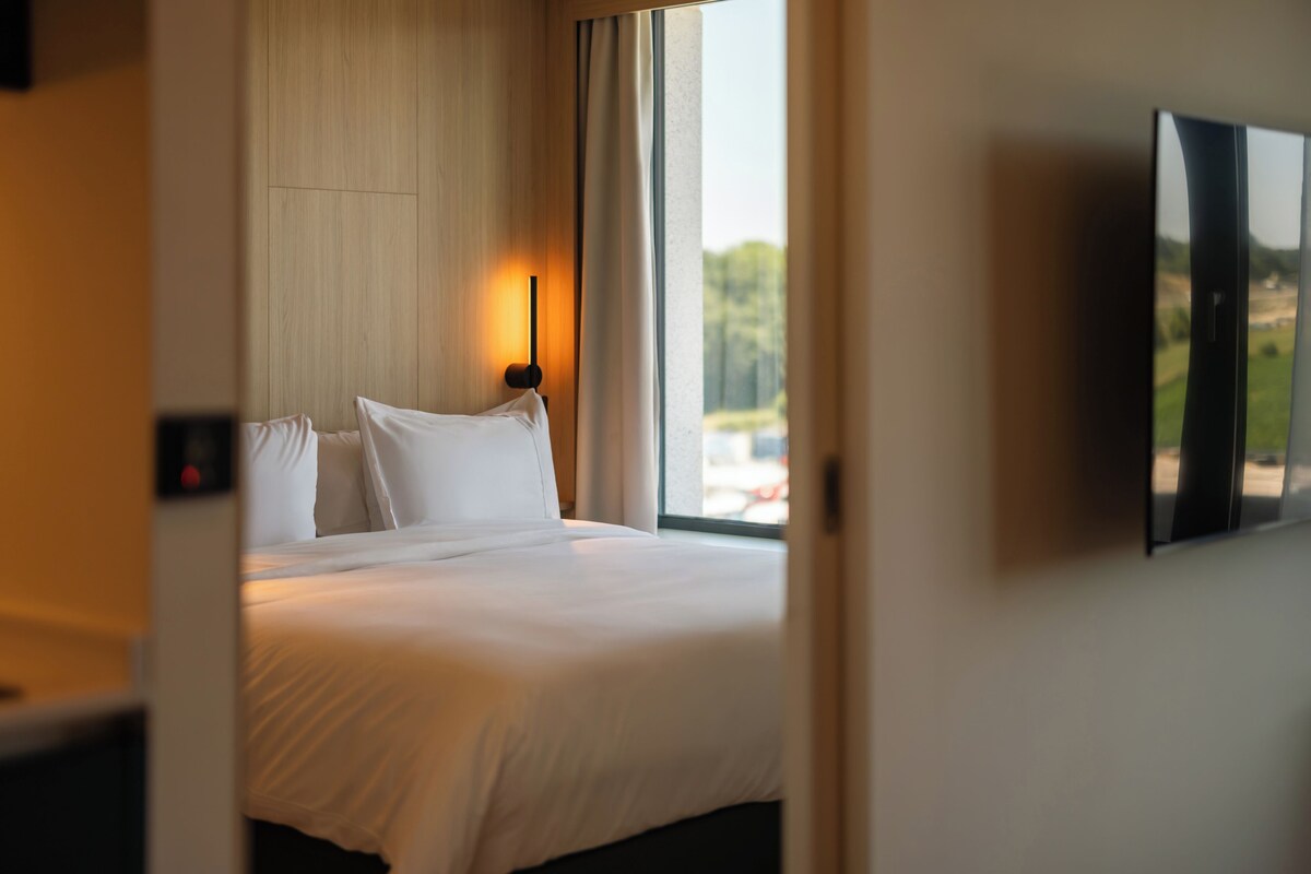 Une chambre d’hôtel avec un lit, un téléviseur fixé au mur et une fenêtre donnant sur un paysage verdoyant.