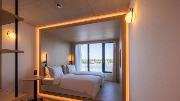 A modern bedroom with a large bed, a small table, and a view of the outdoors through a large window. - NOTO Hotel (Reckange-sur-Mess)