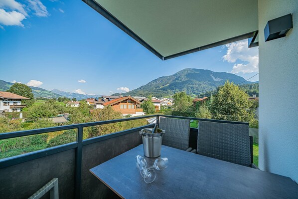 A balcony with a table, a wine glass, and a bottle of wine. The balcony overlooks a mountainous landscape with houses.
