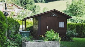 Eine Holzhütte mit geneigtem Dach, einem Fenster und einer Tür, umgeben von einem Garten mit einem Steingartenbeet und verschiedenen Pflanzen.