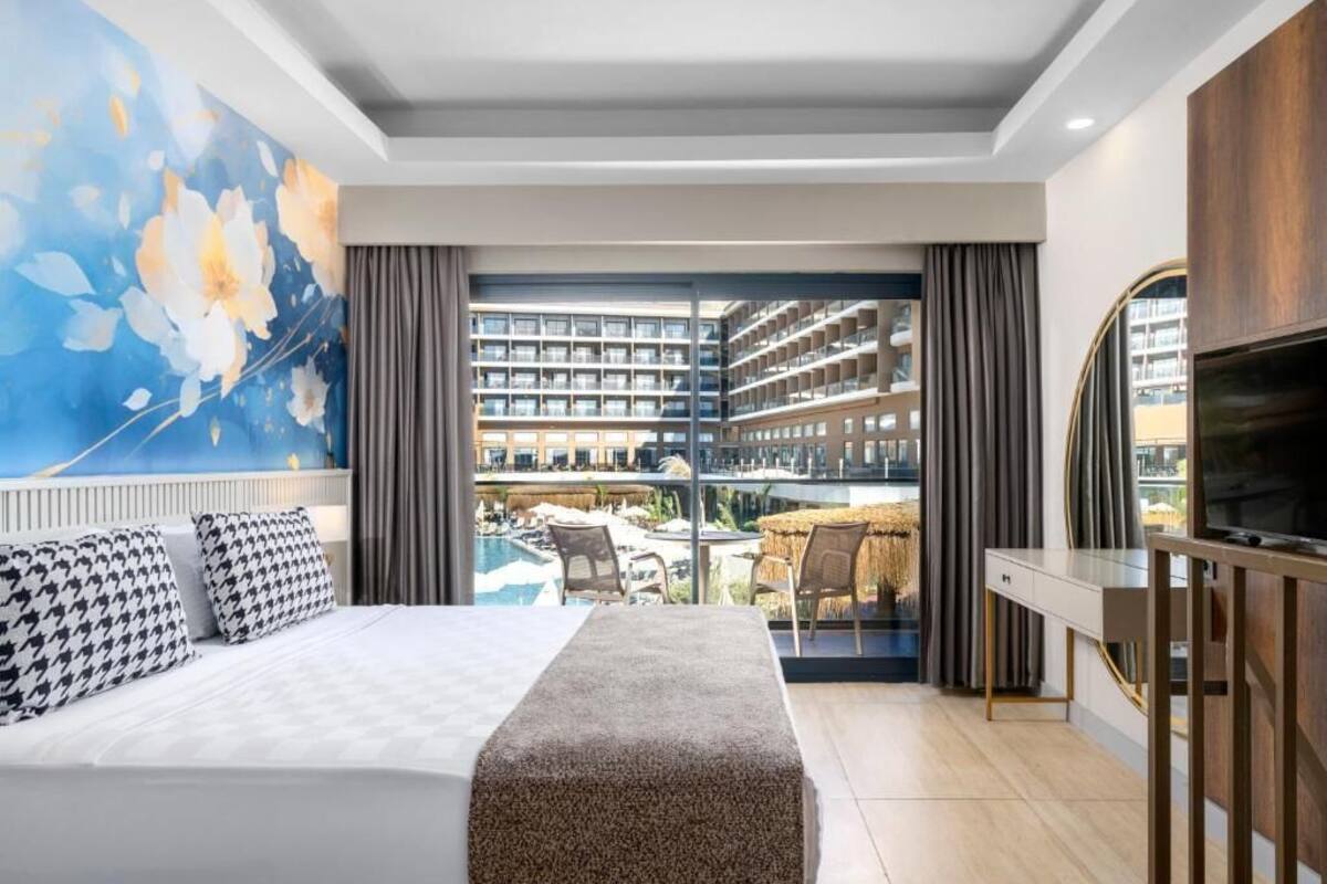 Een hotelkamer met een bed, een televisie, een groot raam met uitzicht op een zwembad en gebouwen, en een schilderij aan de muur.