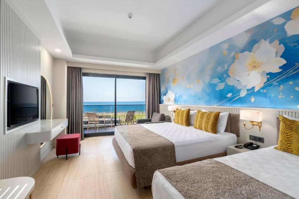 Een hotelkamer met twee bedden, een televisie, een balkon met uitzicht op zee en een grote bloemenmuur.