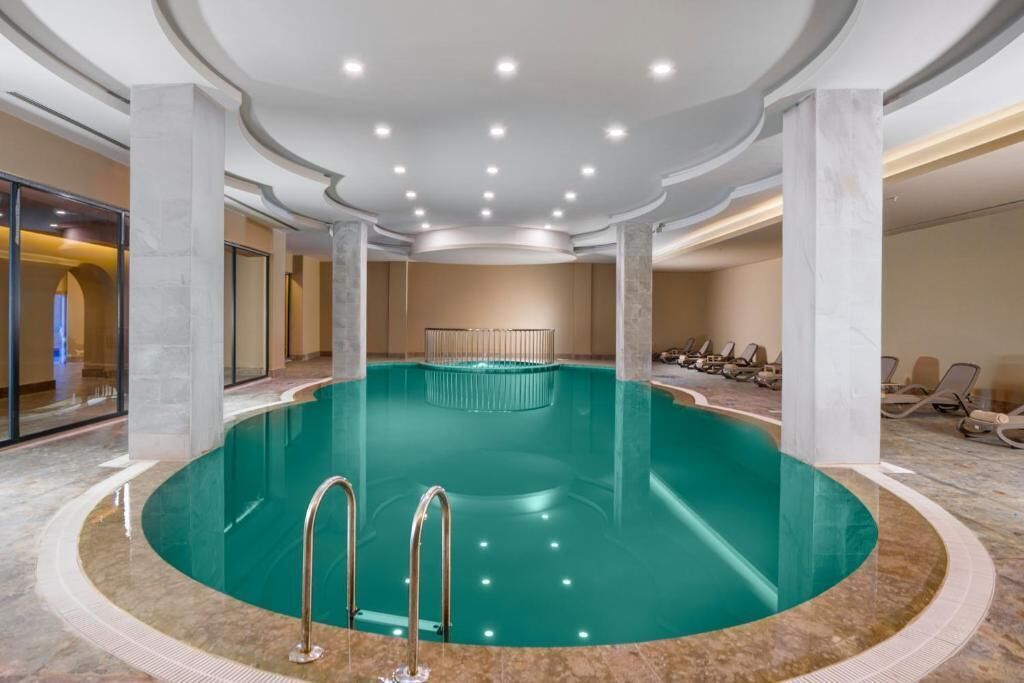 En inomhuspool med en cirkelformad design, omgiven av marmorgolv och pelare, och utrustad med metallräcken.