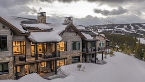 Una gran casa de piedra de varios pisos con techo nevado y múltiples balcones, rodeada de un paisaje nevado y montañas al fondo.