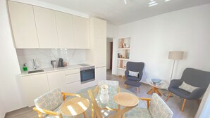 Une cuisine moderne avec des armoires blanches, un four encastré et une machine à café. On y trouve un coin repas avec une table en verre et des chaises à motifs. Un fauteuil gris et une petite table d’appoint avec une lampe sont également visibles.