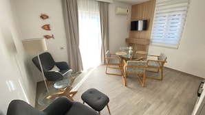 Un salon moderne avec une table basse en verre, deux fauteuils, un canapé et une table à manger avec des chaises. Un téléviseur à écran plat est fixé au mur, il y a un ventilateur de plafond et une fenêtre avec des stores.