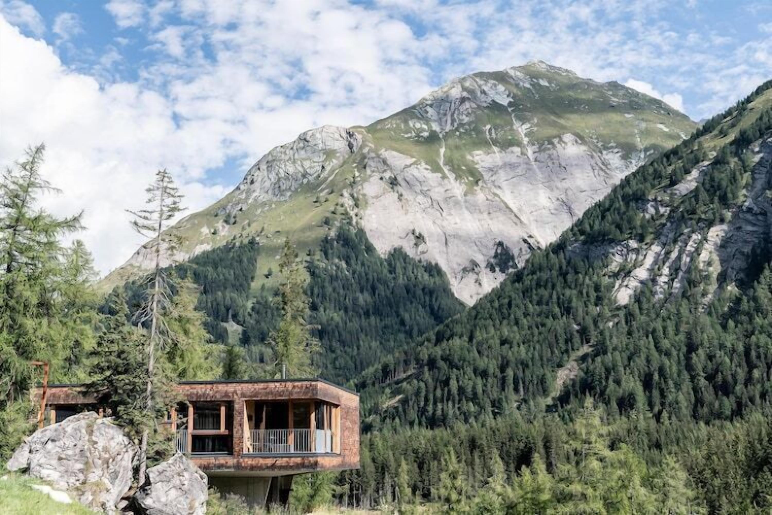 Een berghut met balkon, omringd door dicht bos en een grote berg op de achtergrond.