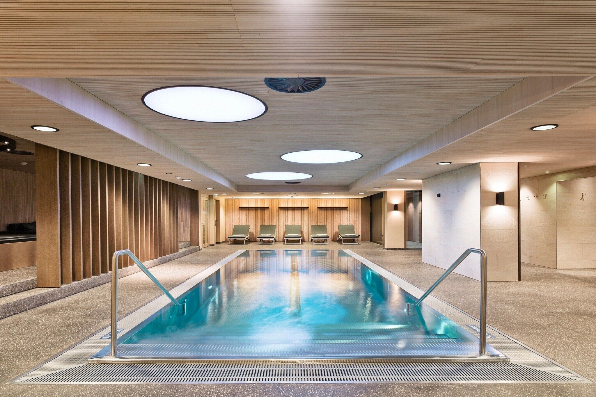 A modern indoor swimming pool with a clear water surface, surrounded by lounge chairs and a wooden panel wall.