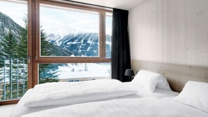 A bedroom with a large window offering a mountain view, a wooden headboard, and white bedding. - Chalet Resort near Lake (Kals am Großglockner)