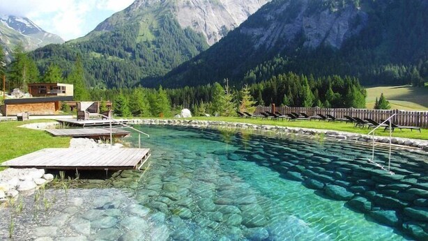 A clear pool with a wooden deck, surrounded by a wooden fence and trees, with mountains in the background. - Chalet Resort near Lake (Kals am Großglockner)