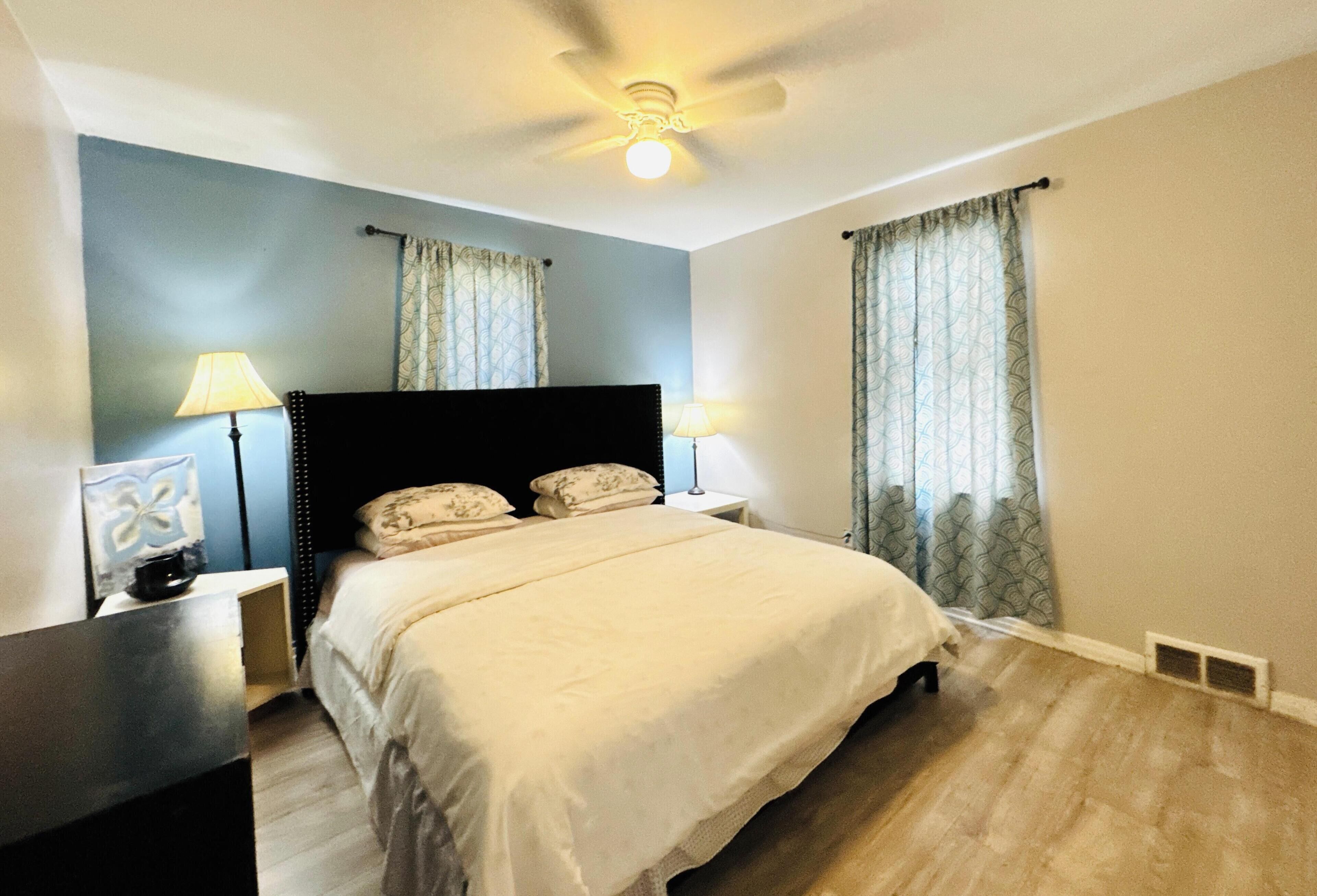 A bedroom with a bed, two bedside lamps, a ceiling fan, and curtains.