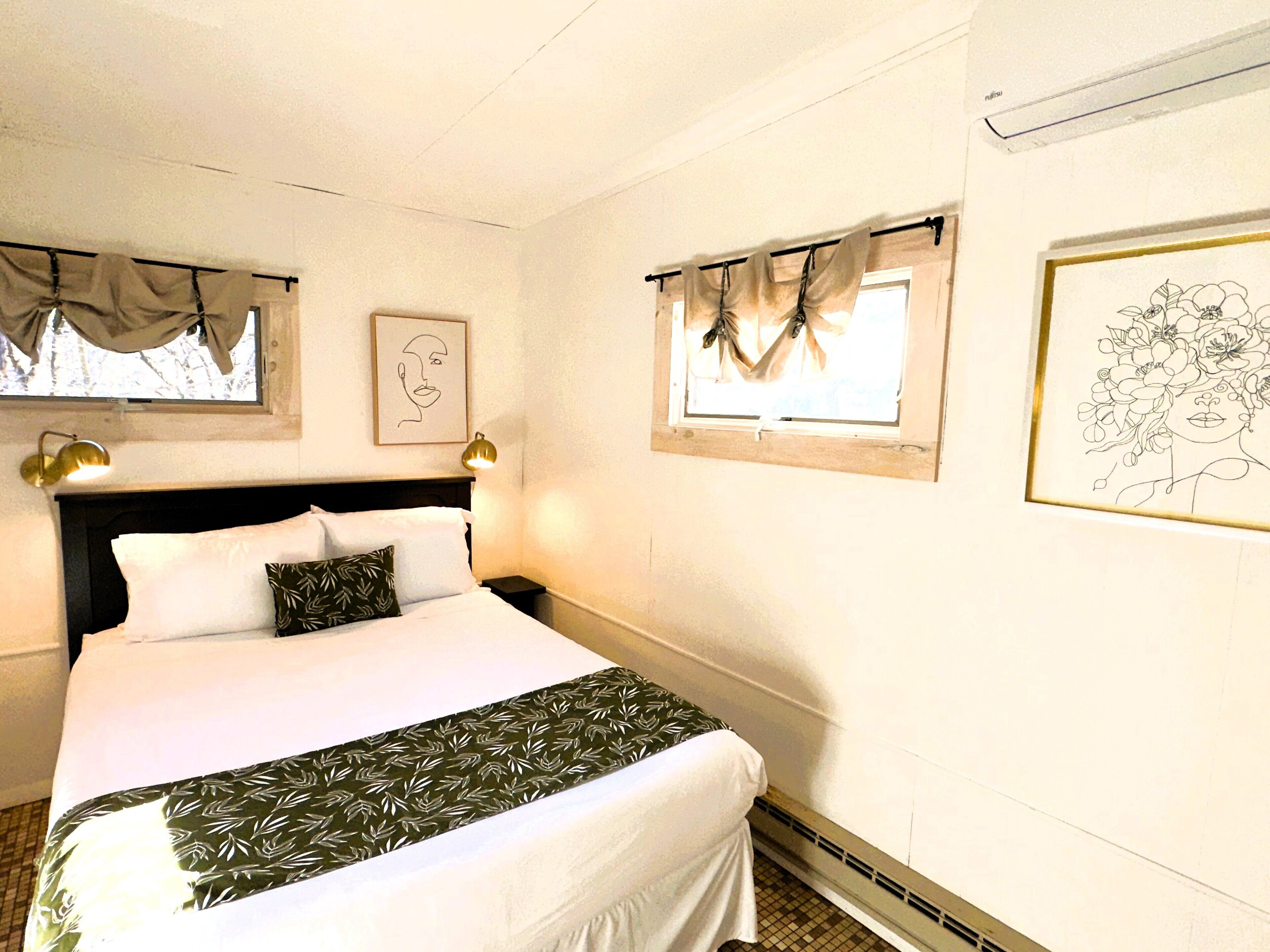 A neatly made bed with a patterned bedspread, a headboard, and two bedside lamps. There are two windows with curtains, one with a framed picture on the wall, and a wall-mounted air conditioner.