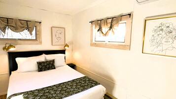 A neatly made bed with a patterned bedspread, a headboard, and two bedside lamps. There are two windows with curtains, one with a framed picture on the wall, and a wall-mounted air conditioner.