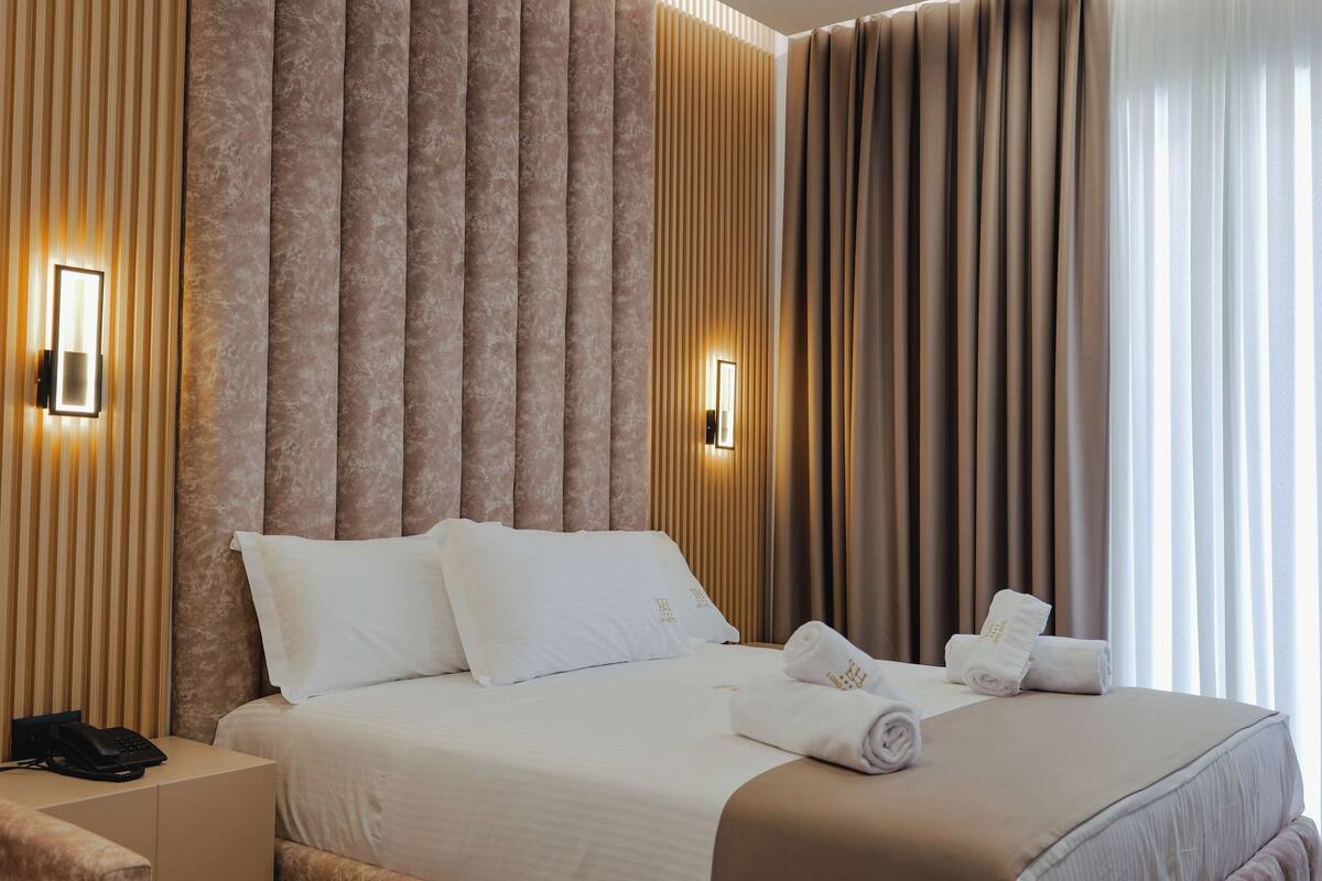 Habitación de hotel con una cama grande, dos mesitas de noche, un teléfono en la pared y una ventana con cortinas.