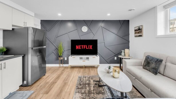 A modern living room with a flat-screen TV displaying 'NETFLIX', a white sofa, a coffee table, and a kitchen area with a refrigerator and cabinets. - Your Comfort Spot in Niagara |Mins away from Falls (Niagara Falls)