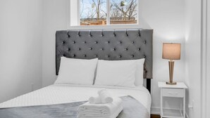 A neatly made bed with white linens and a grey tufted headboard, a bedside lamp, and a window with a view of bare trees. - Your Comfort Spot in Niagara |Mins away from Falls (Niagara Falls)