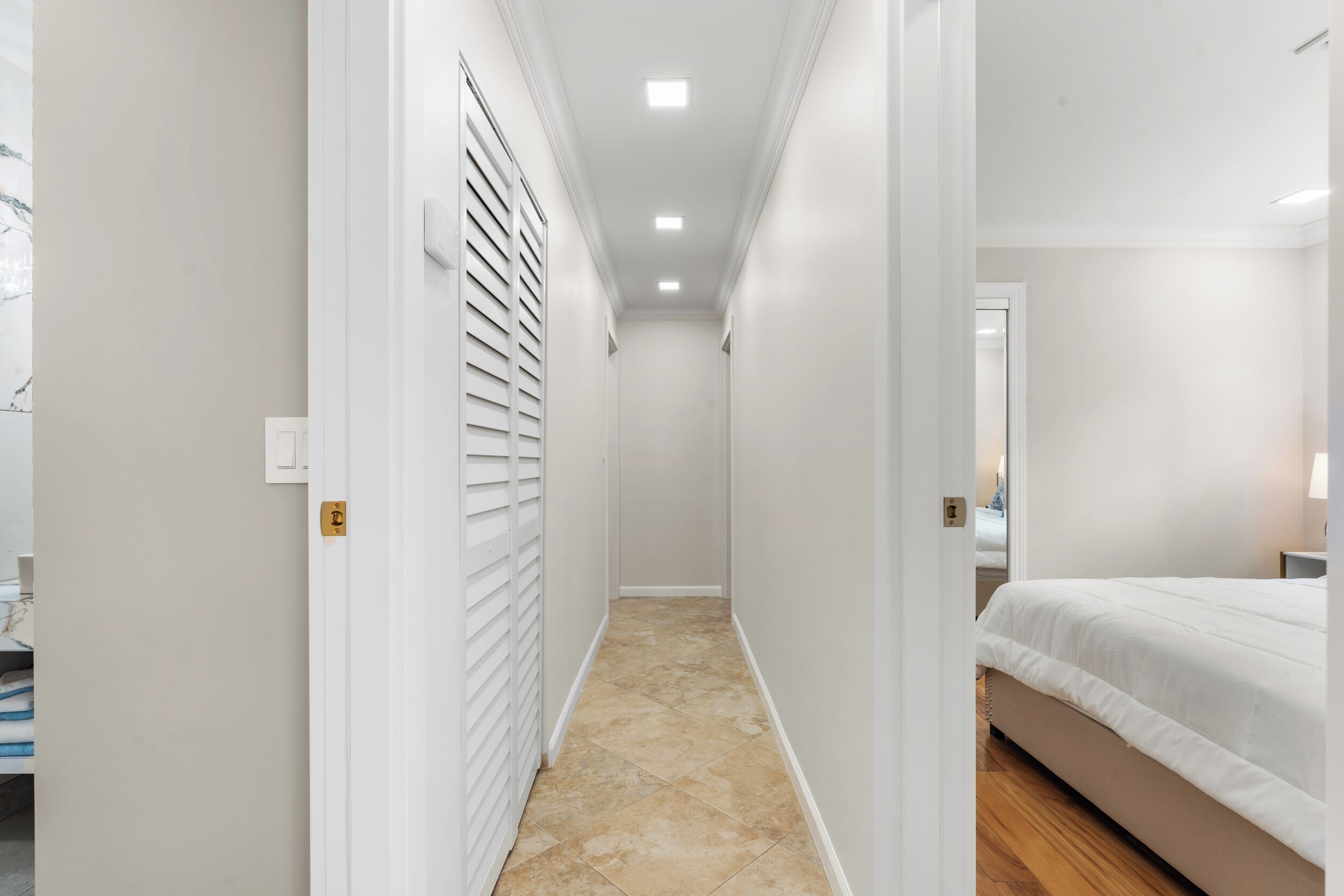 A narrow hallway with a bed on the right, a door with a lock on the left, and a tiled floor.