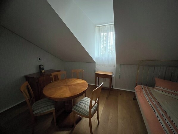 Een kamer met een houten eettafel en stoelen, een bed met beddengoed met een patroon, een radiator en een raam met dunne gordijnen.