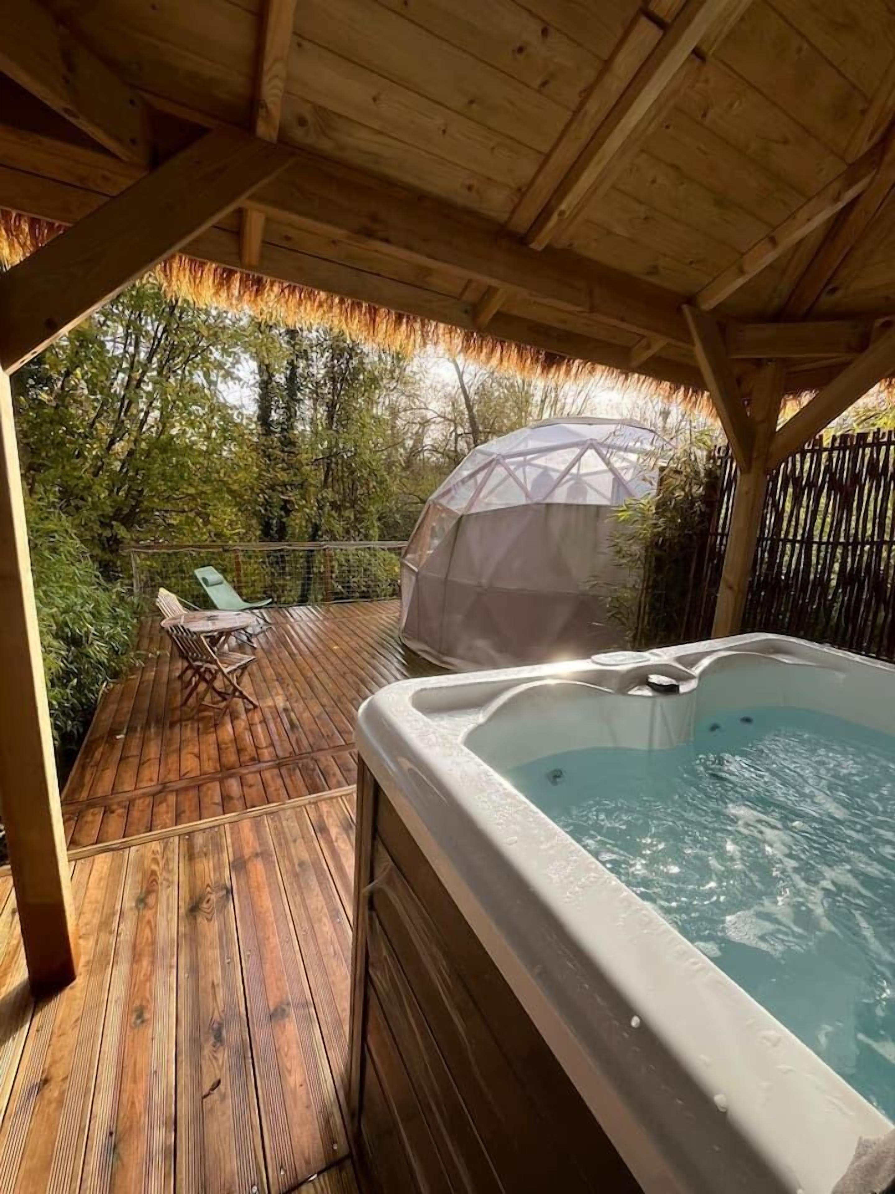 Une terrasse en bois avec un jacuzzi, des chaises longues et une structure recouverte d’un toit de chaume.