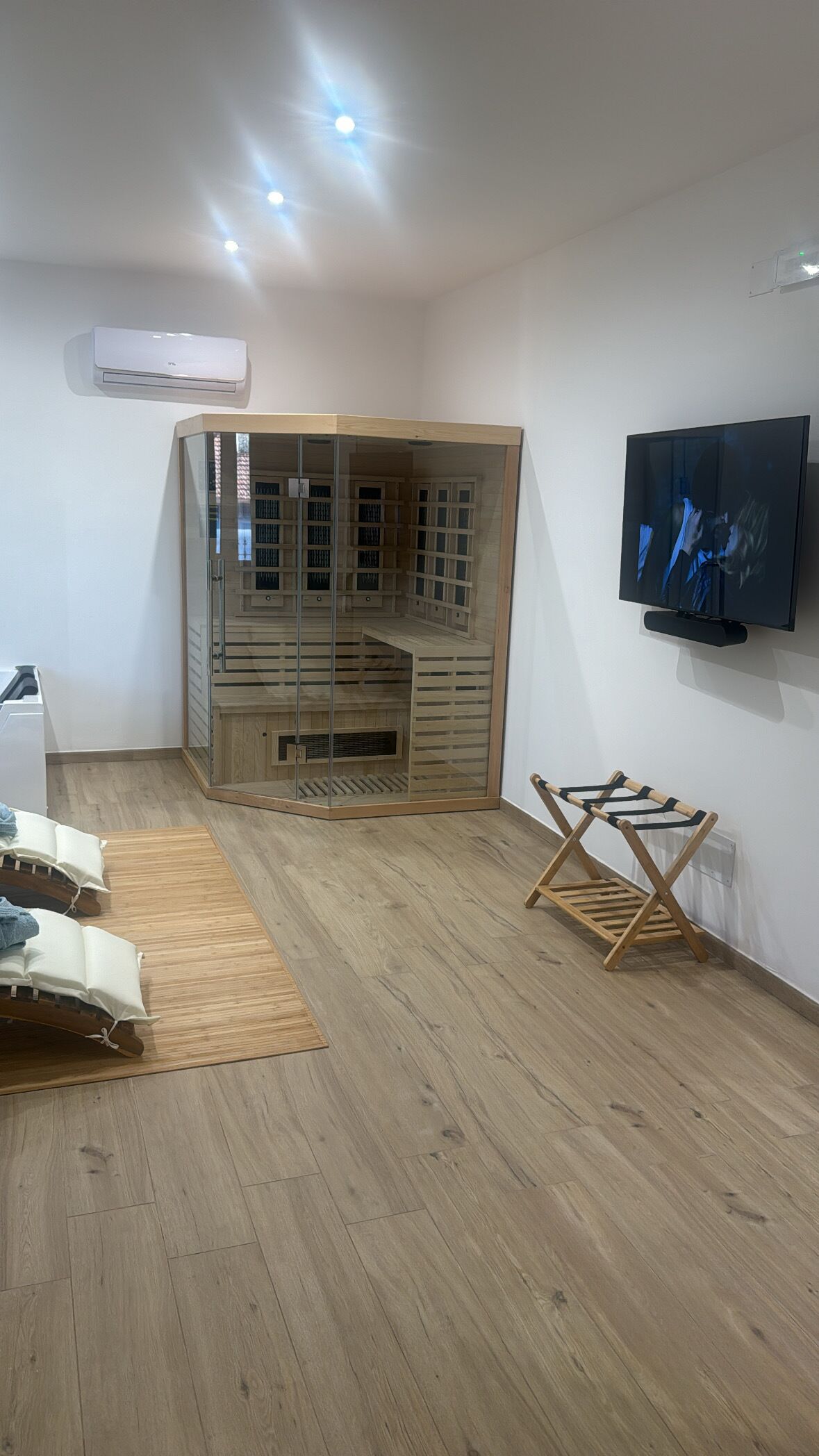A room with a wooden floor, a glass partitioned seating area, a mounted TV, and a ceiling-mounted air conditioner.