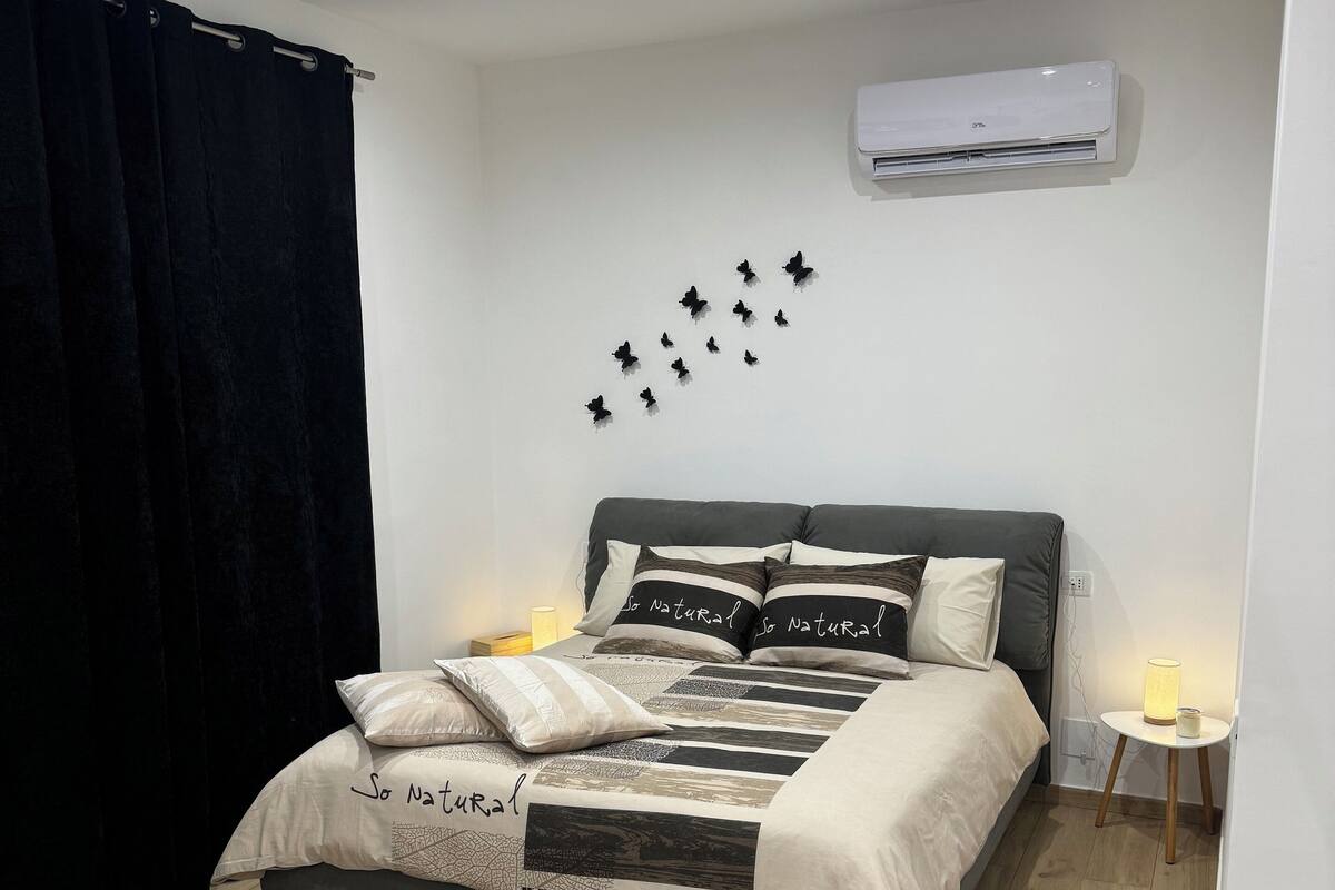 A bedroom with a bed, a wall-mounted air conditioner, a bedside table with a lamp, and a black curtain.