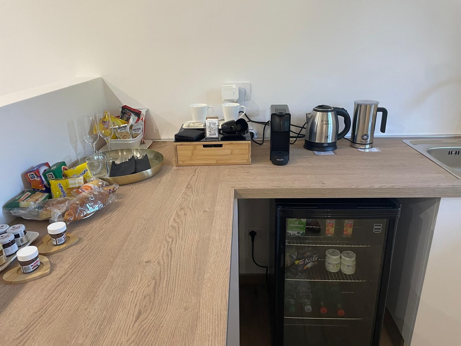 A compact kitchen with a built-in refrigerator, a coffee maker, a kettle, and various food items on the counter.