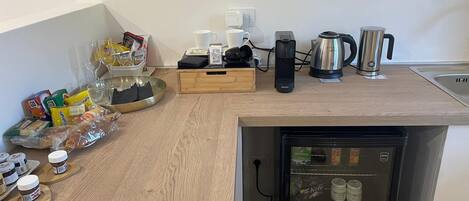 A compact kitchen with a built-in refrigerator, a coffee maker, a kettle, and various food items on the counter.