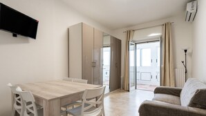 Una sala moderna con una mesa de comedor de madera, sillas blancas, un sofá beige y un televisor de pantalla plana montado en la pared.