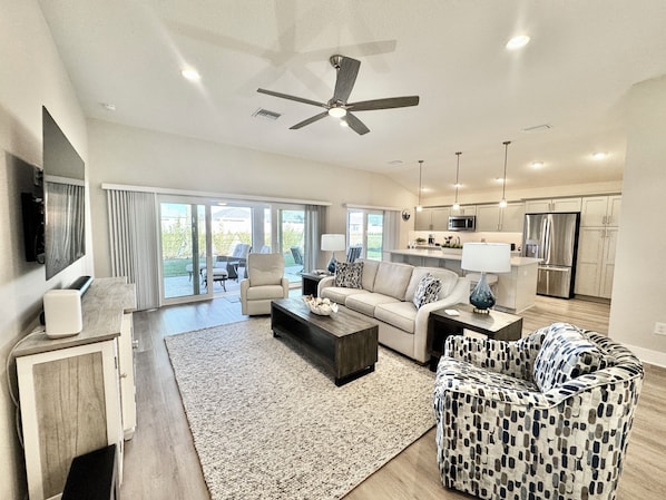A spacious living room with a sofa, coffee table, and patterned ottoman. There is a kitchen area with modern appliances and a ceiling fan.