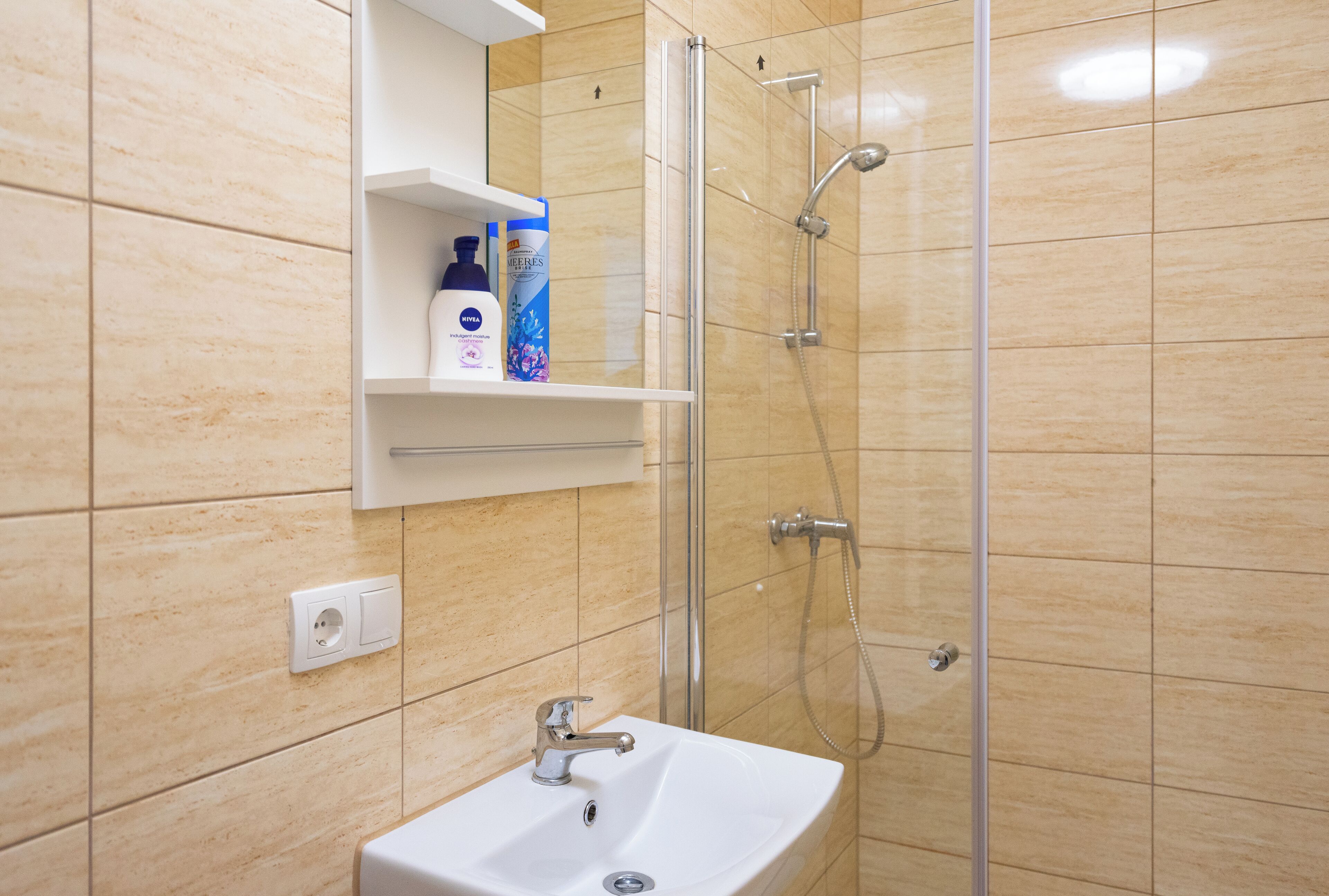 Un baño con lavabo, cabina de ducha de cristal y estante con artículos de aseo.