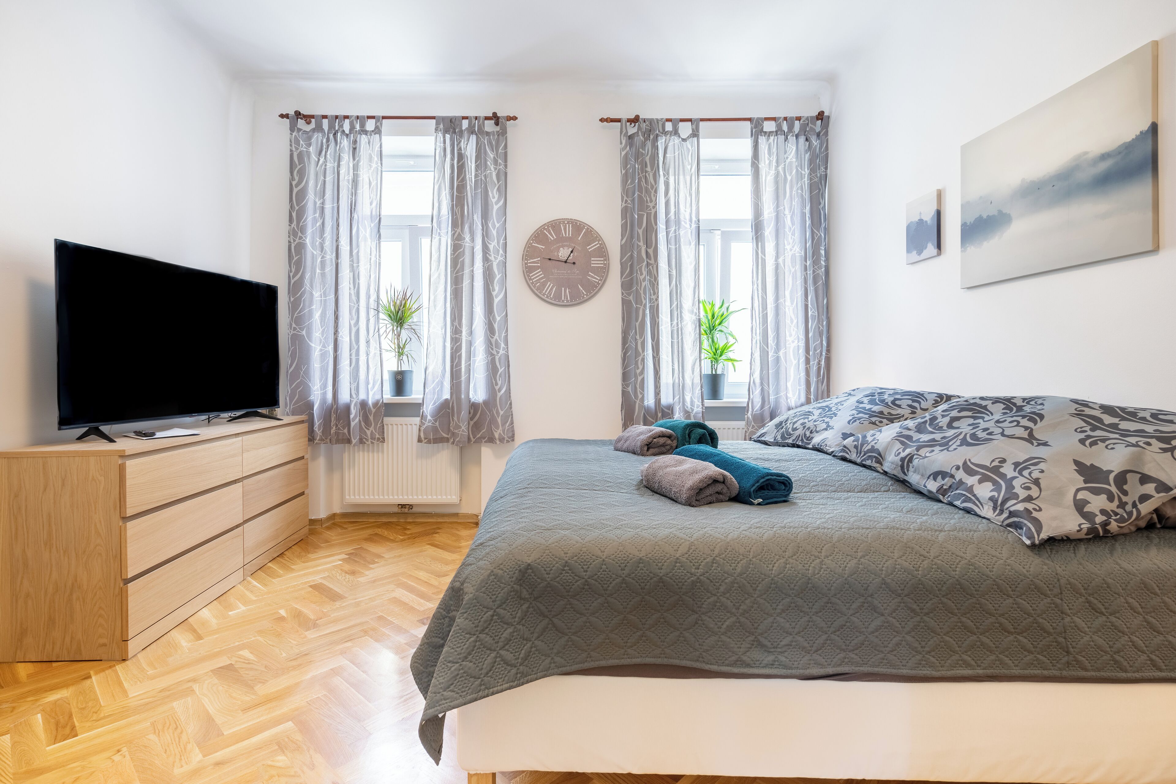 Un dormitorio con una cama, un televisor sobre un aparador de madera, dos ventanas con cortinas, un reloj y un cuadro en la pared.