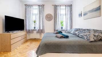 Un dormitorio con una cama, un televisor sobre un aparador de madera, dos ventanas con cortinas, un reloj y un cuadro en la pared.
