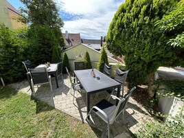 A patio with a table and chairs, surrounded by greenery and a house in the background.