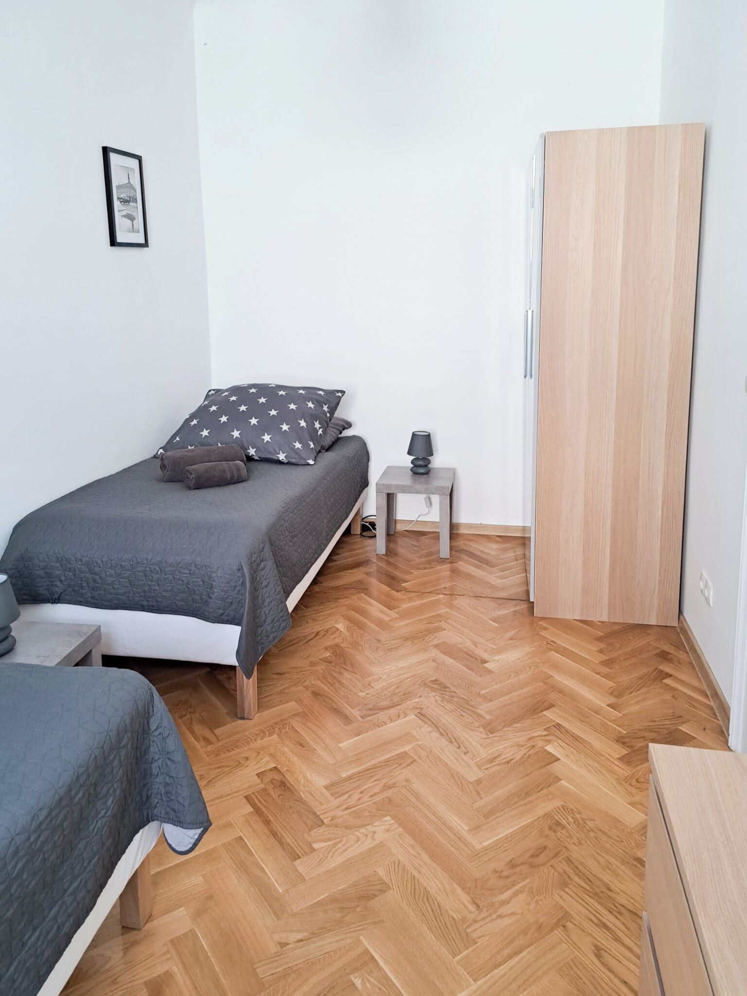 A bedroom with a bed, a bedside table, a lamp, a wardrobe, and a herringbone-patterned wooden floor.
