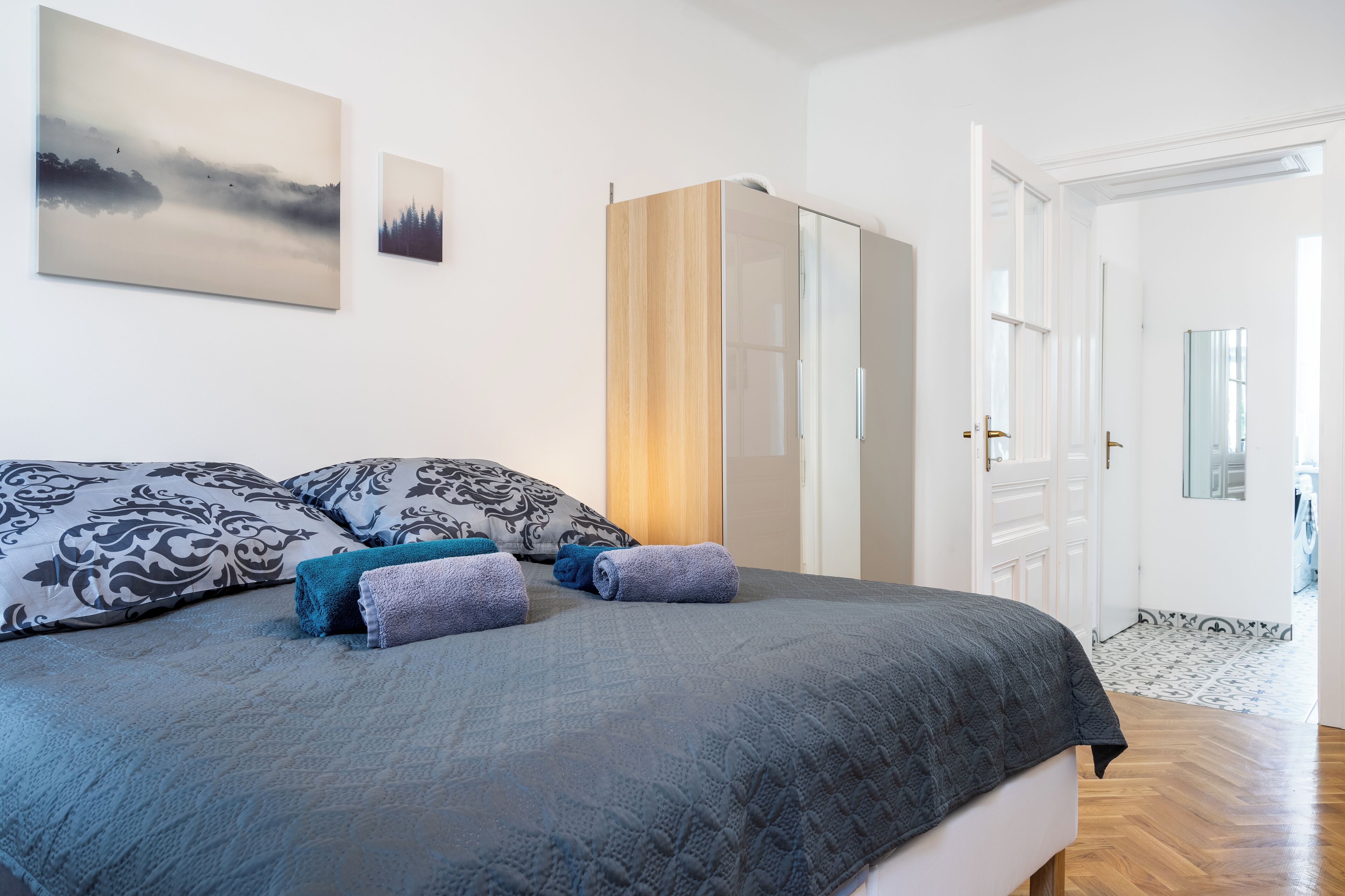 A neatly made bed with a grey quilt, two pillows with a black and white pattern, and two rolled towels in blue and purple.