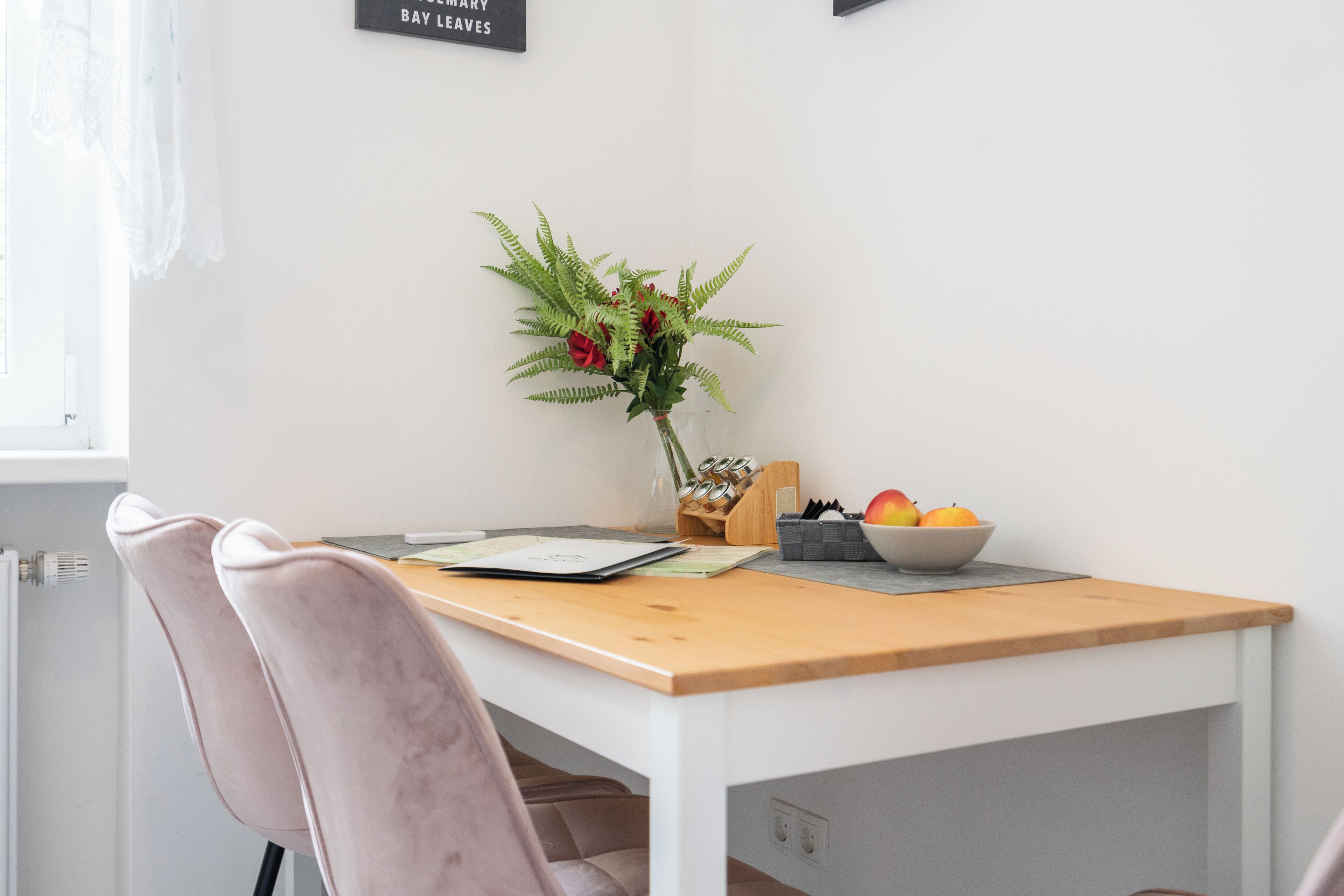 Un comedor con una mesa blanca, dos sillas rosadas, un jarrón con flores rojas y follaje verde, un frutero y un letrero en la pared.