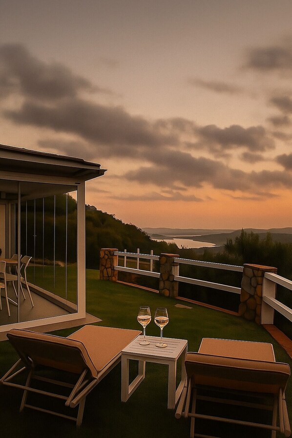 A patio with outdoor furniture and a table with two wine glasses, overlooking a scenic landscape with hills and a body of water.