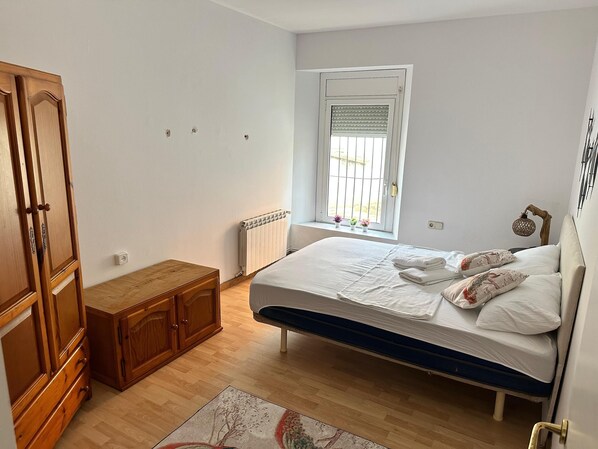 A bedroom with a bed, wooden wardrobe, radiator, window with blinds, and a small table. - Vista Alegre Rest House 8 Person (Vallromanes)