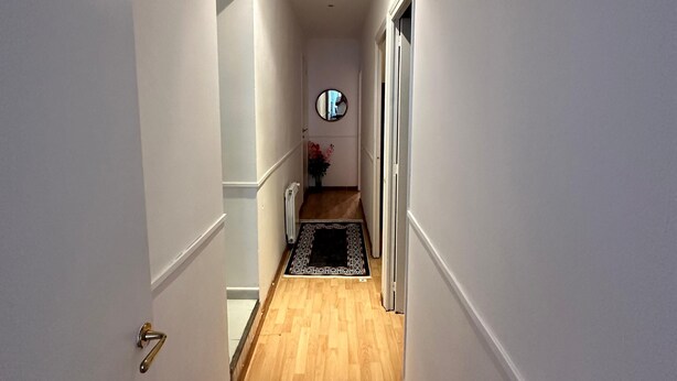 Un pasillo estrecho con suelo de madera, paredes blancas y un tapete blanco y negro que lleva a una habitación con un espejo redondo en la pared.