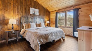 Un dormitorio de cabaña de madera acogedor con una cama, dos mesitas de noche con lámparas, un ventilador de techo y un gran ventanal con cortinas.