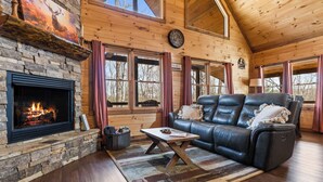 Interior acogedor de una cabaña con chimenea de piedra, sofá de cuero y ventanas amplias.