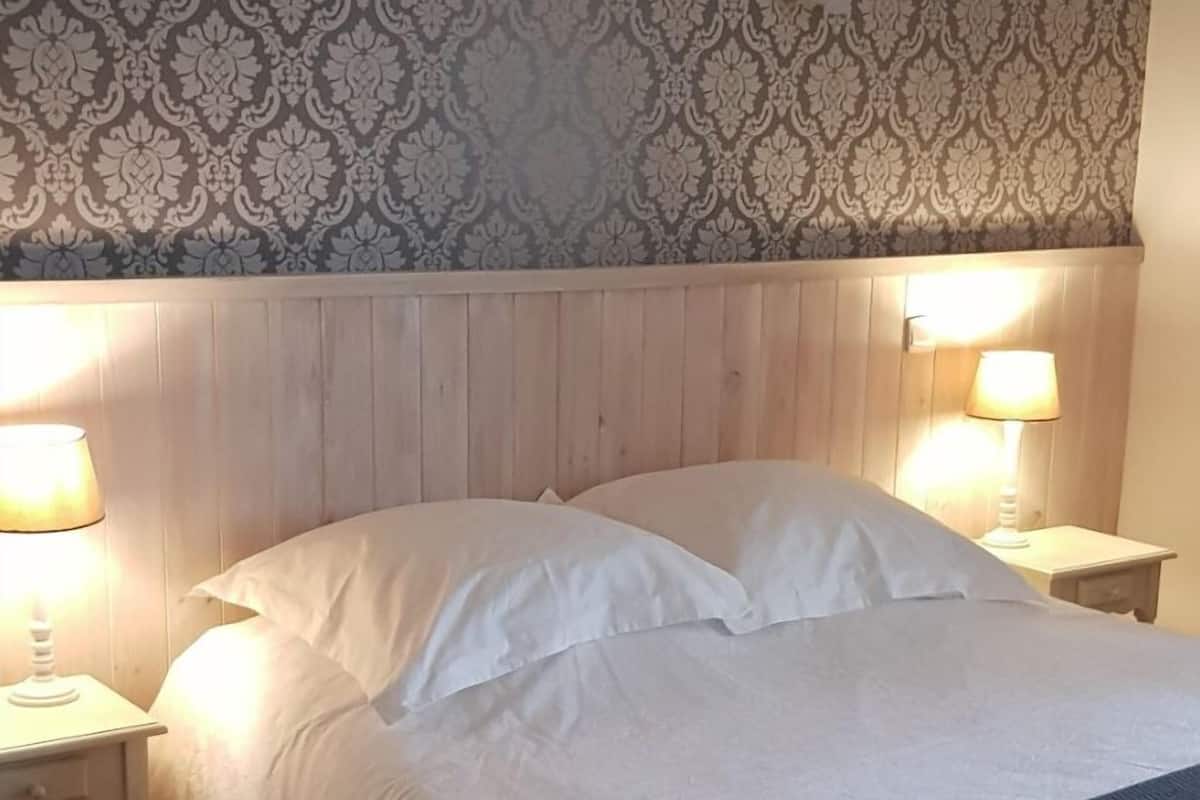 A bedroom with a wooden headboard, a bed with white bedding, two bedside lamps, and a patterned wallpaper.