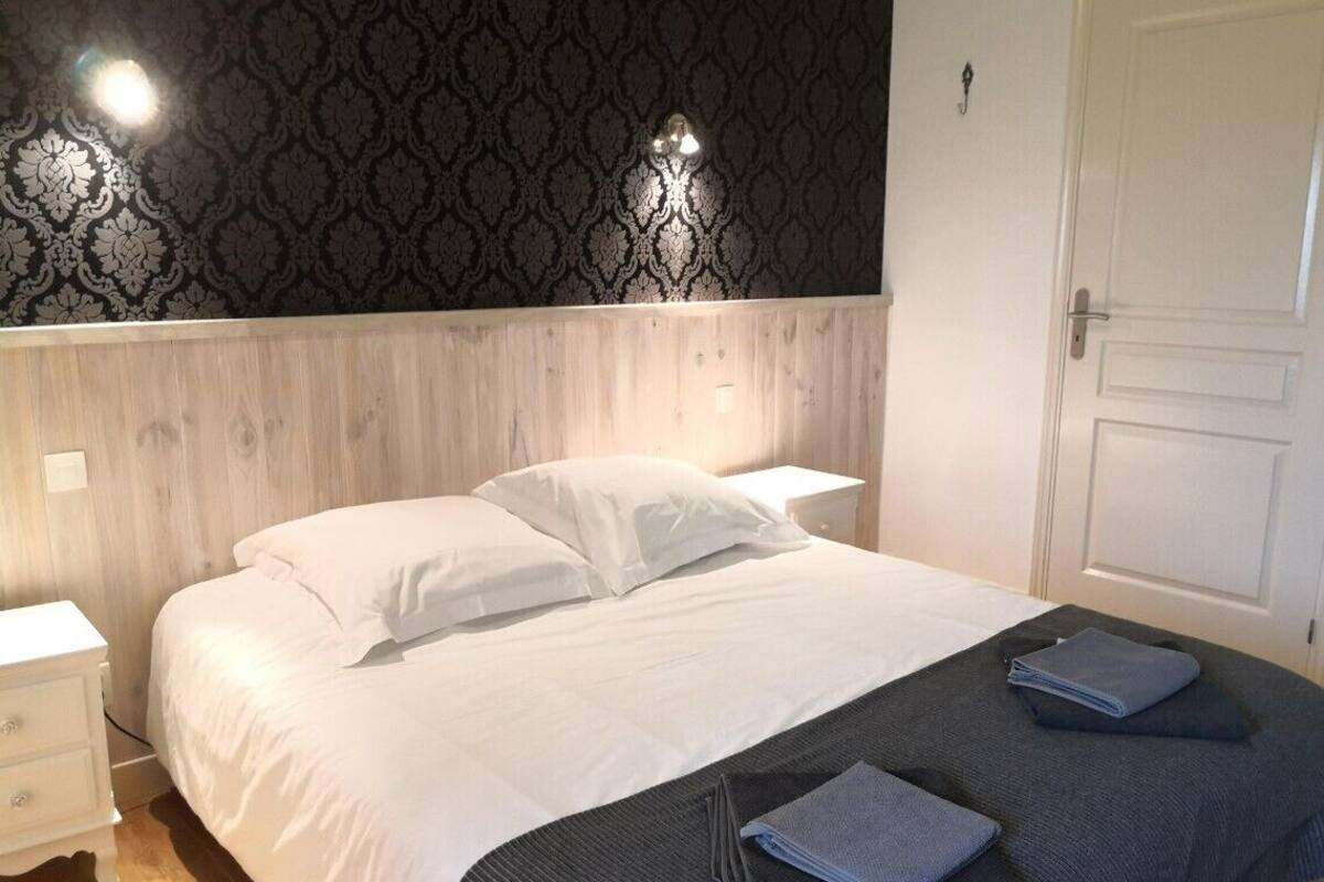 A neatly made bed with white linens and a dark gray throw blanket, a bedside table with a lamp, and a patterned wallpaper on the wall.